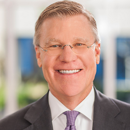 Smiling man with gray hair and glasses in a dark suit and purple tie