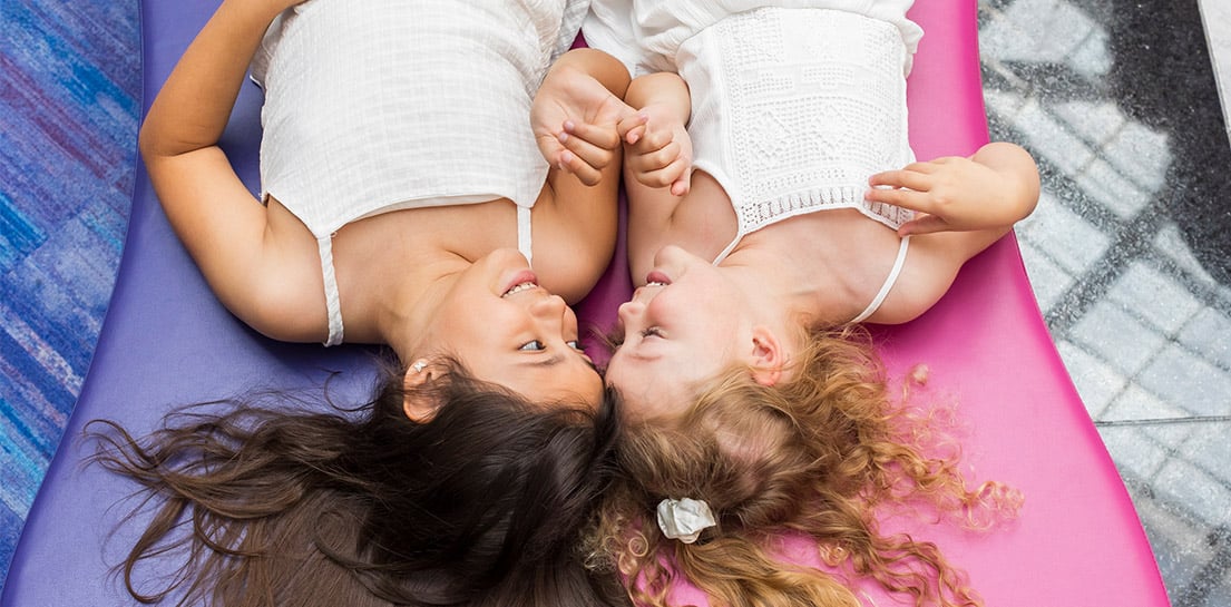 Two smiling girls lie heads together, holding hands. One has dark hair, the other blonde