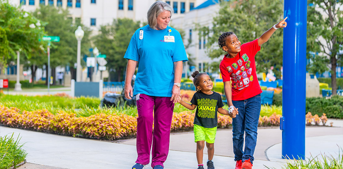 Woman in scrubs holds hands with two smiling children. One in a SAVAGE shirt, the other points upwards