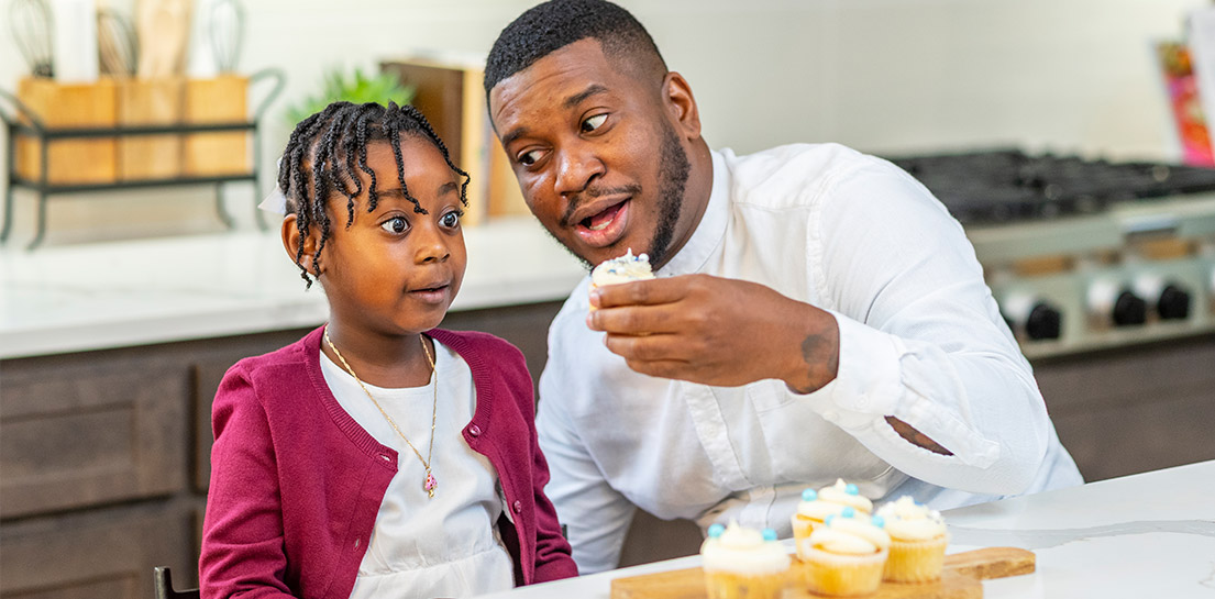 Man playfully offers cupcake to wide-eyed girl. More cupcakes on a kitchen counter