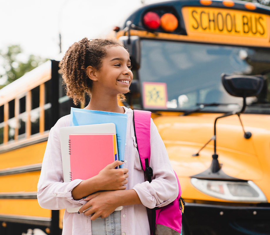 African American girl holding school notebooks and standing next to yellow school bus