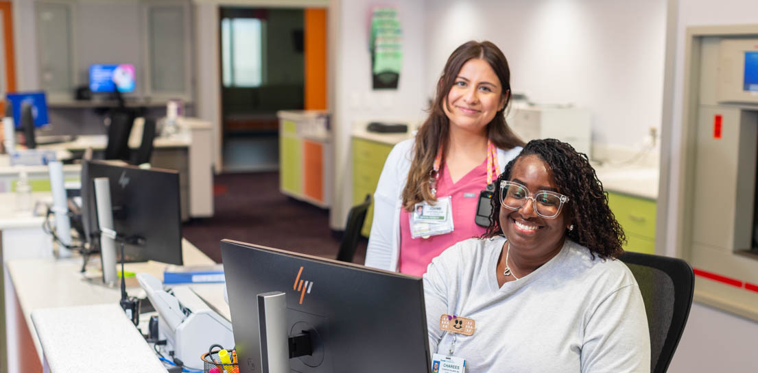 Nurse standing behind a trainee on a computer smiling