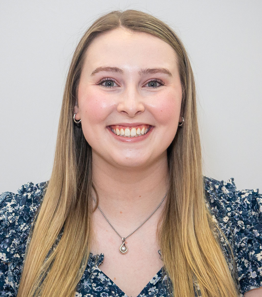 Smiling young woman with blonde hair, light eyes, wearing a floral top and pearl pendant necklace