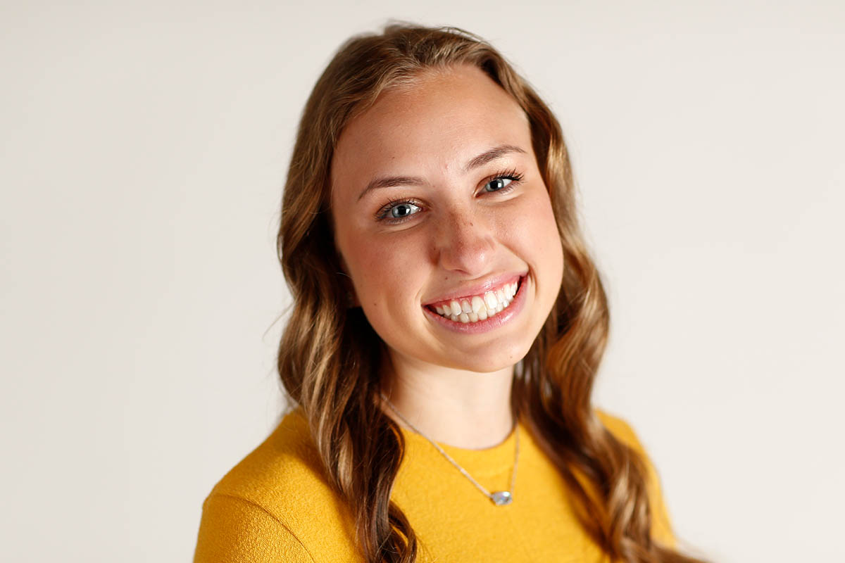 Smiling young woman with long brown hair and blue eyes, wearing a yellow shirt and silver necklace