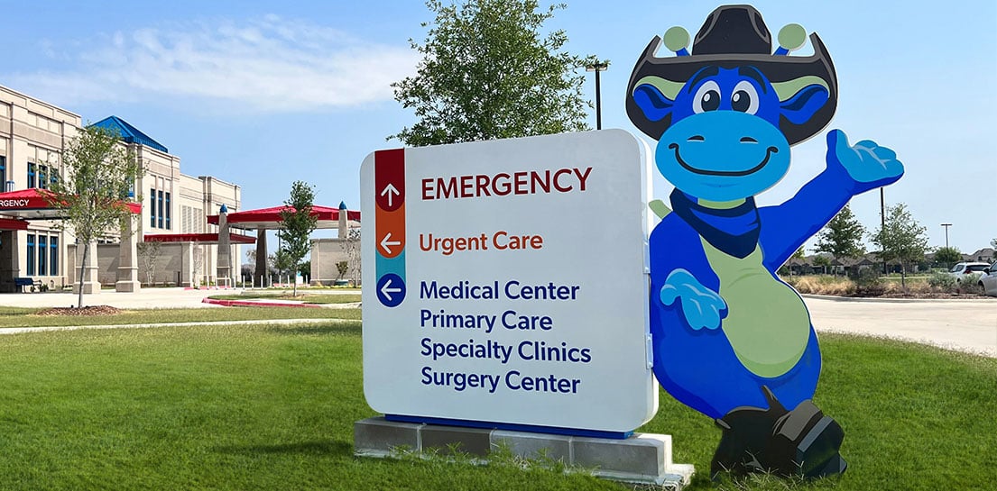 Blue dinosaur mascot by sign: EMERGENCY (↑), Urgent Care (←), Medical Center (←), Primary Care, Specialty Clinics, Surgery Center