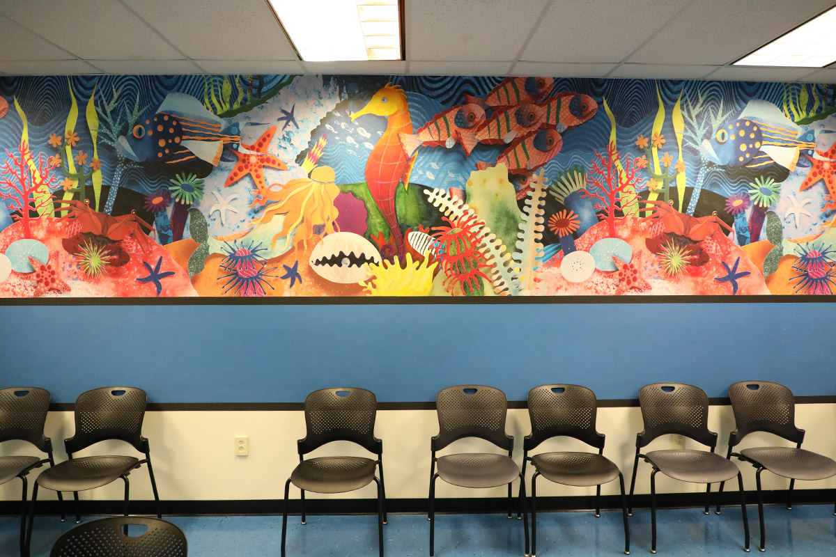 Colorful mural of an undersea scene with fish, coral, and a seahorse above a row of black chairs in a waiting room
