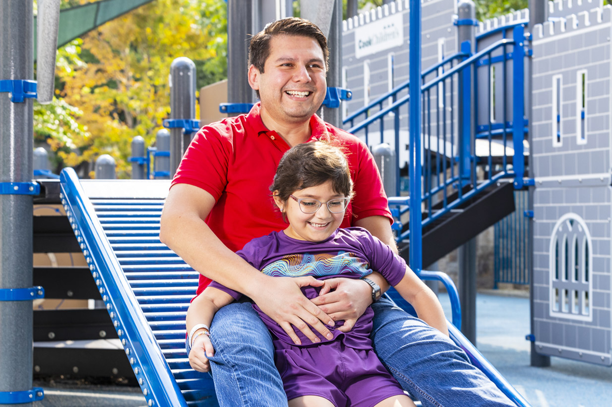 Man and child smiling while sliding down a blue playground slide; Cook Children's on a structure