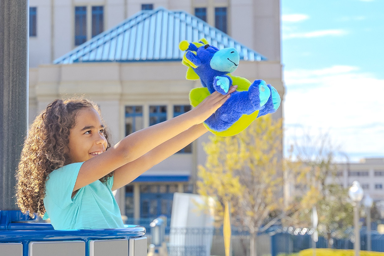 Smiling girl with curly hair holds a blue and green plush dragon toy up to the sky