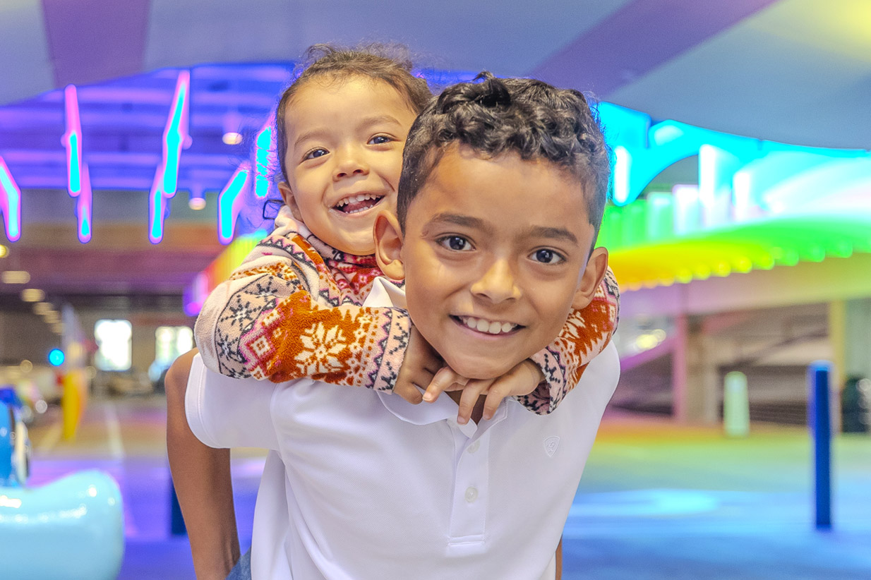 Two smiling children, an older boy piggybacking a younger girl, in a colorful indoor setting