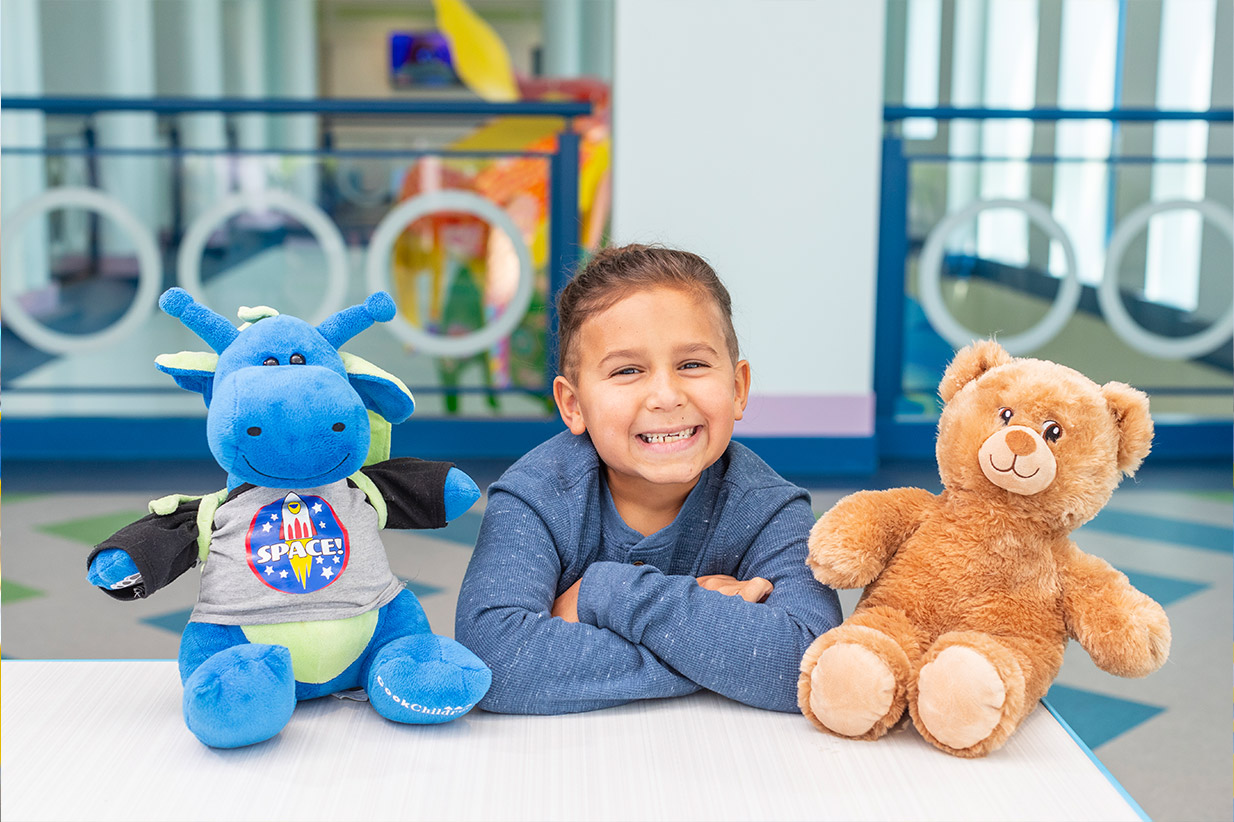 Smiling child with two stuffed animals, one blue with a SPACE! shirt, and one a brown bear