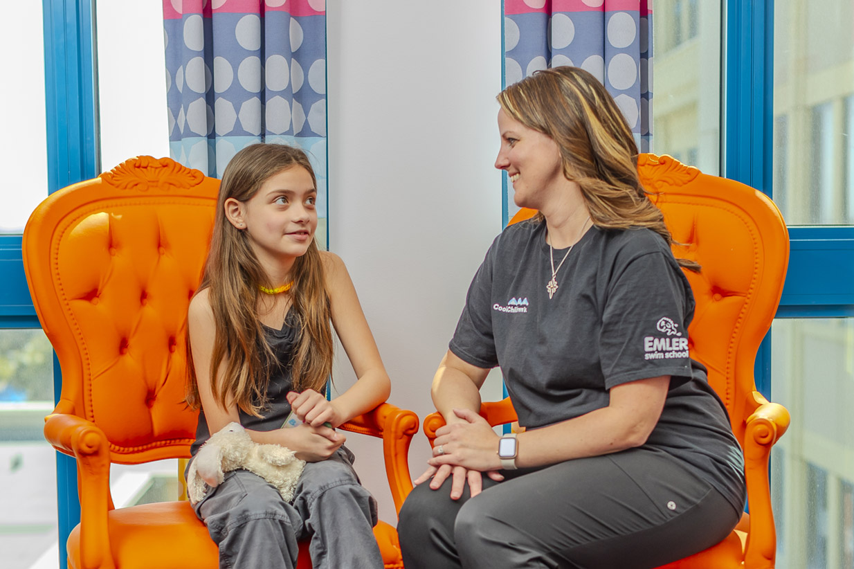 Woman with CookChildren's and EMLER swim school logos on shirt talks to a girl while sitting in orange chairs