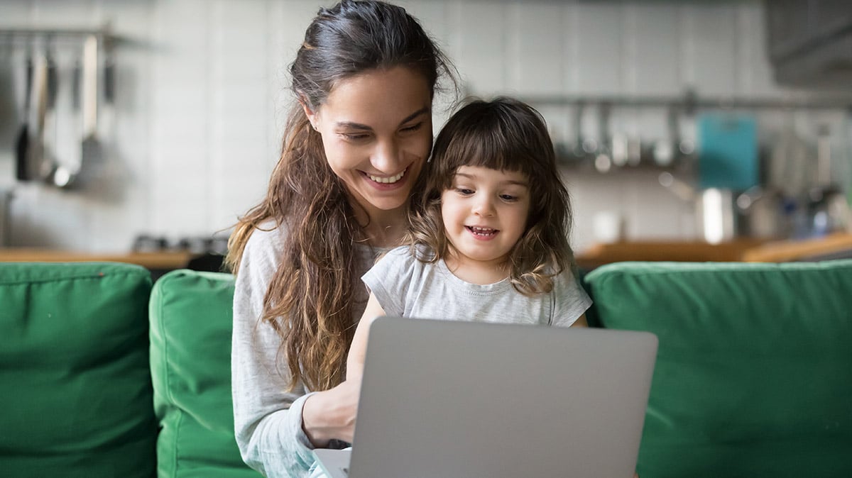 Smiling mother and young child happily look at a laptop together on a green couch