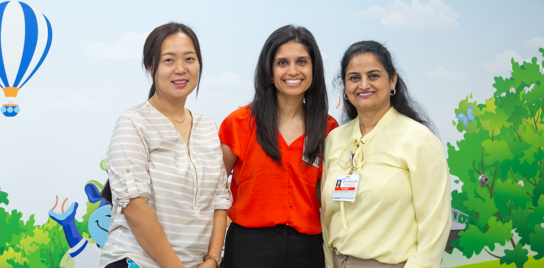 Three smiling women stand in a clinic with a whimsical mural. One woman's ID badge reads DR KALLUR