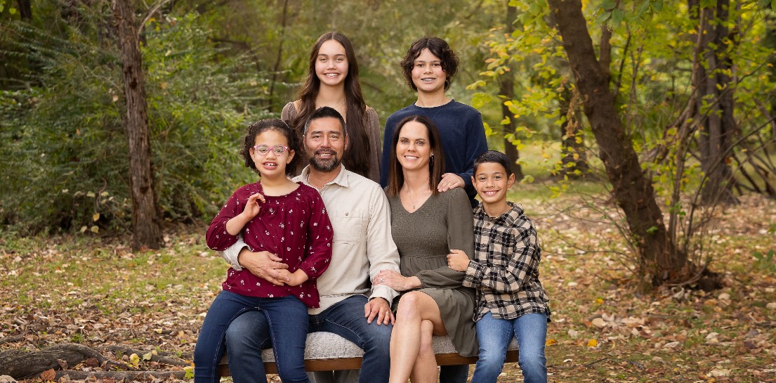 A smiling family of six: two adults and four children, on a bench in a park with autumn trees
