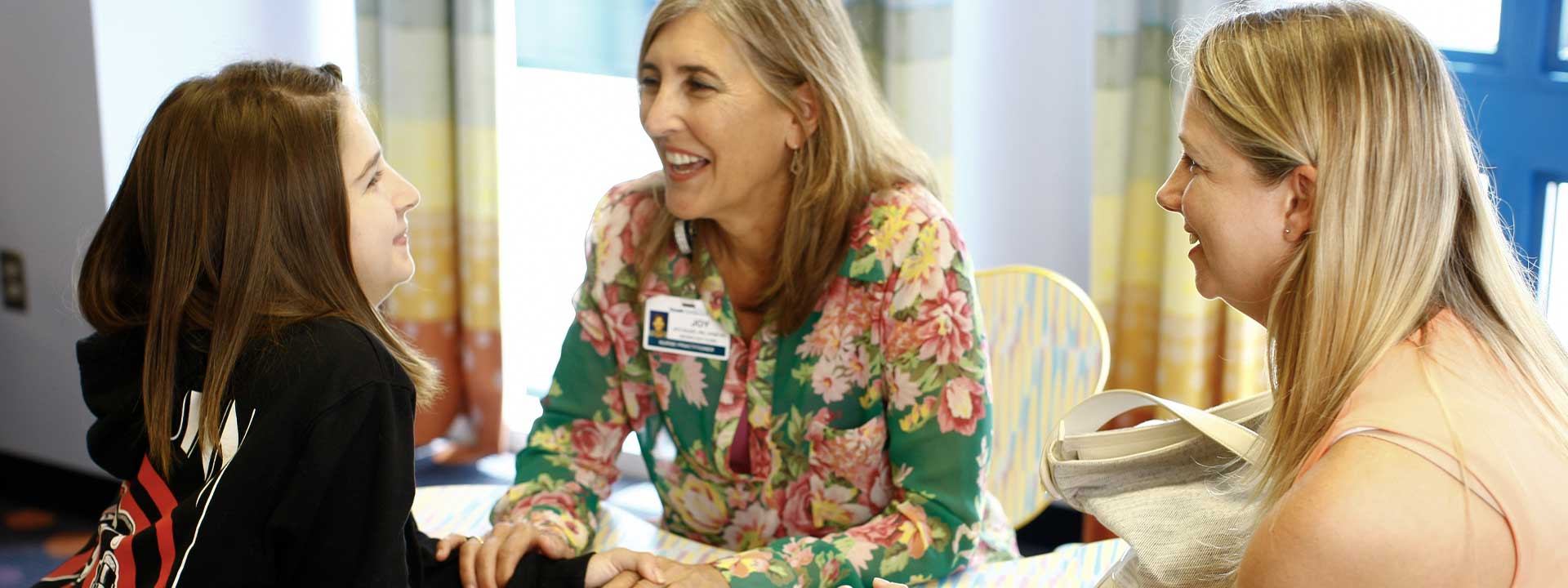Three women smiling and talking. One wears a nametag: JOY, FAMILY NURSE PRACTITIONER