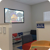 Children's clinic waiting area with a TV showing a cartoon, seating, and a 'WELL CHILD WAITING AREA' sign