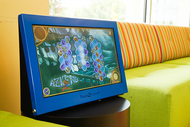 Interactive screen with game 'Azkend 2'. A hand cursor points to a hexagonal tile. Text: Touch2PLAY kids