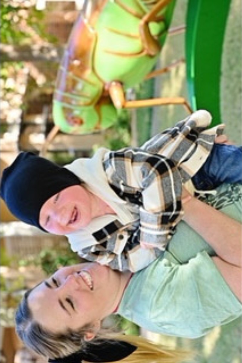 Woman holds laughing baby in beanie and plaid shirt; large green grasshopper sculpture behind them