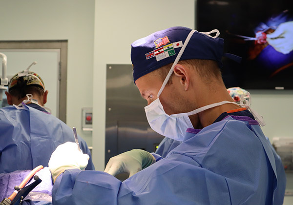 Doctor Hansen in blue scrubs, mask, and cap with country flags operates. Another surgeon in background