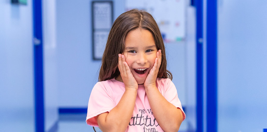 Smiling girl with long brown hair, hands on cheeks, mouth open in surprise and joy in a blue room