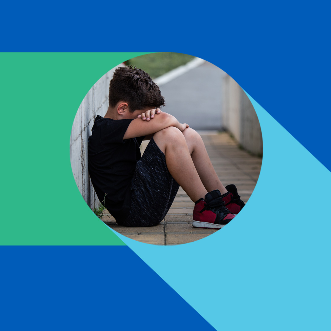 Young boy sits hunched on ground, head in arms, appearing distressed and alone. Circular image with blue and green background.