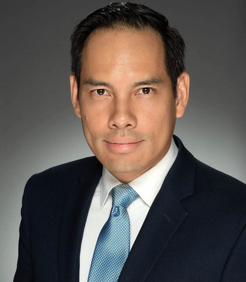 Man in a dark suit and blue tie, looking forward with a slight smile against a grey background