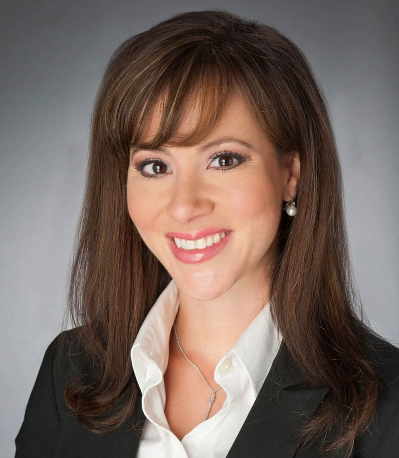 Headshot of a smiling woman with brown hair, white shirt, black blazer, and pearl earrings
