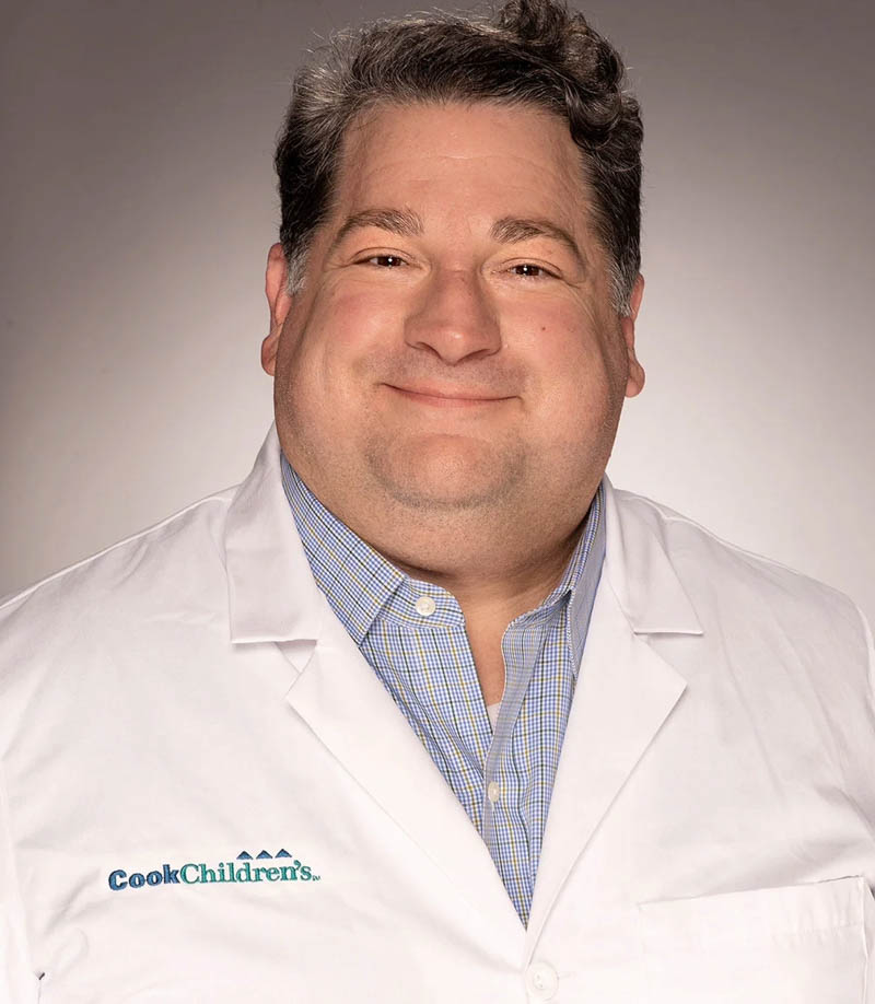 Smiling man in white lab coat and plaid shirt with CookChildren's logo