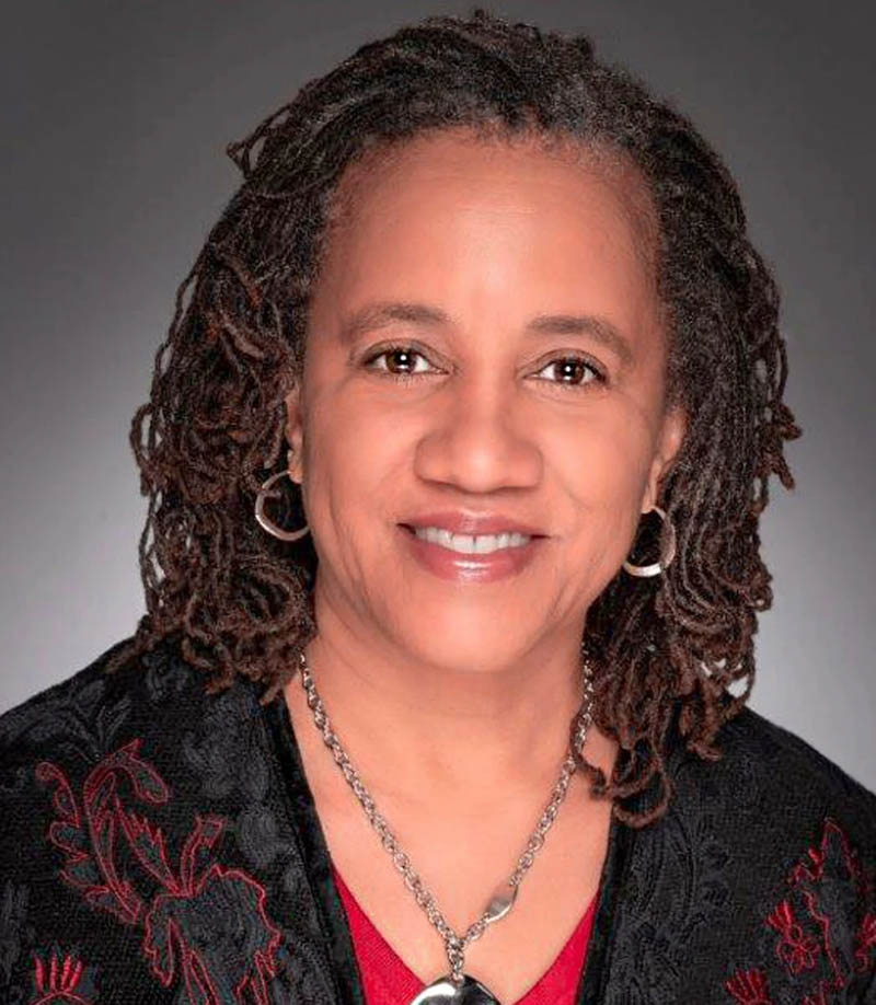 Smiling woman with dreadlocks, wearing a red shirt, black embroidered jacket, silver necklace, and hoop earrings