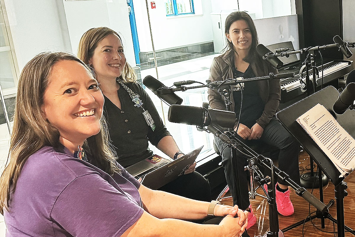 Three smiling women sit at microphones with a laptop, sheet music, and keyboard