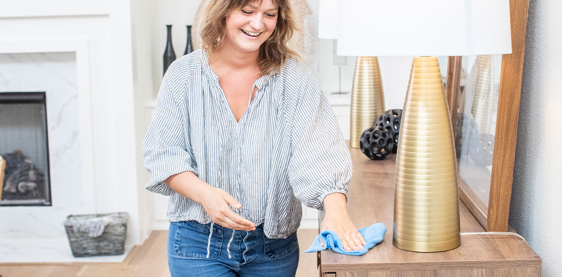 Woman in blue striped blouse and jeans dusting furniture and lamp