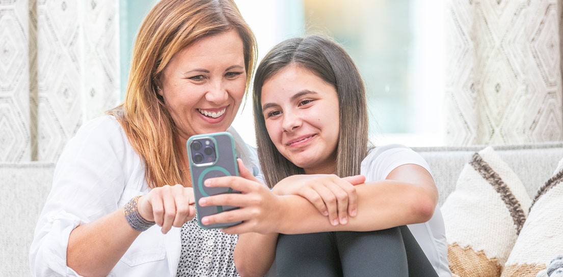 Mother and young teen daughter sitting together looking at mobile phone and smiling