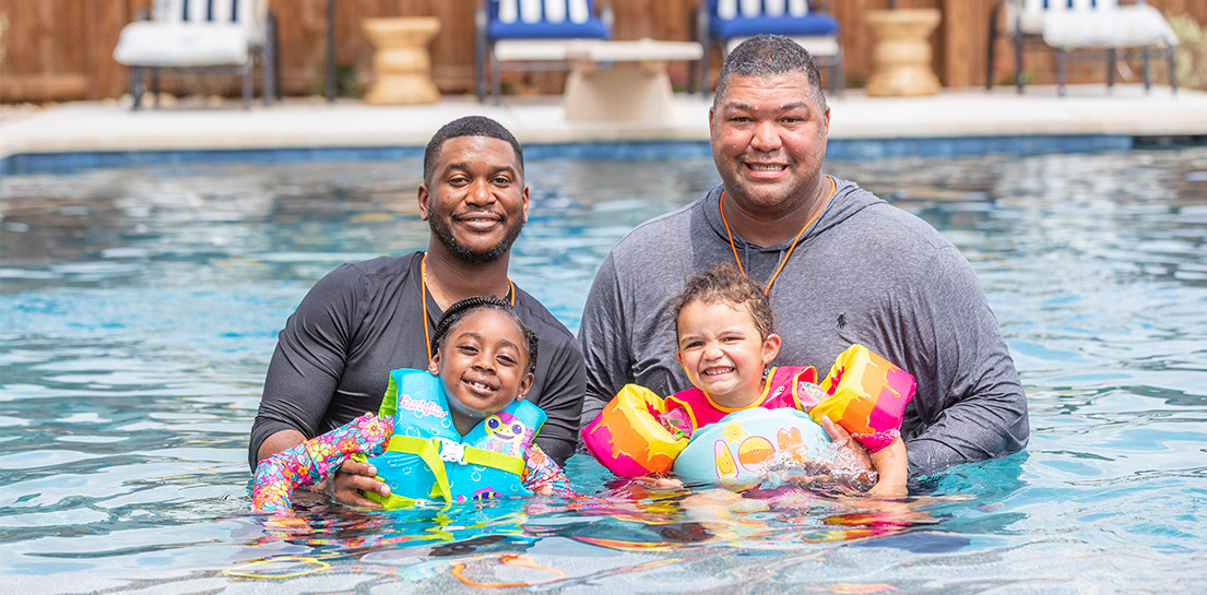 Two dads in swimming pool holding toddlers wearing life jackets