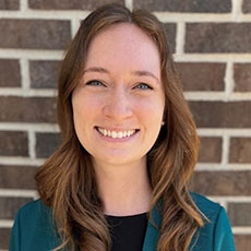 Professional headshot of a smiling young woman with long brown hair against a brick wall