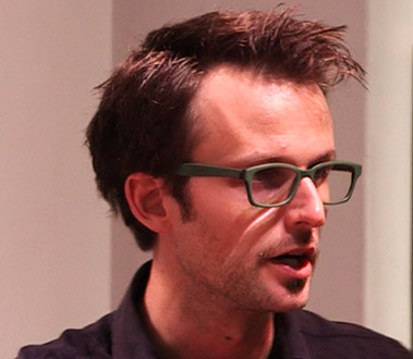 Man with dark hair and green glasses, wearing a dark shirt, speaking