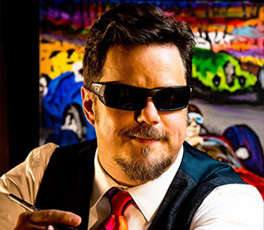 Smiling man in dark sunglasses, vest, and colorful tie, holding a pen. Abstract car paintings in background