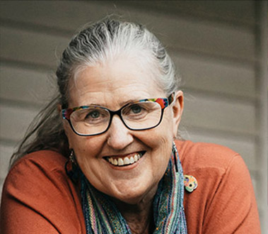 A smiling woman with gray hair and colorful glasses, wearing an orange top and a patterned scarf, looks at the camera