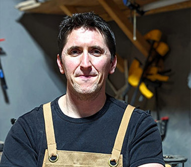 A smiling man in a black shirt and brown apron in a workshop, looking at the camera