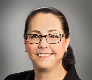 Woman with dark hair, glasses, and pearl earrings smiles