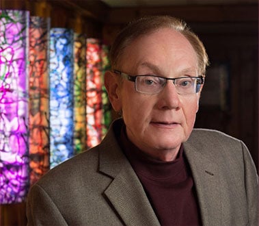 Older man with glasses, gray blazer, and maroon turtleneck, in front of colorful, illuminated art installations