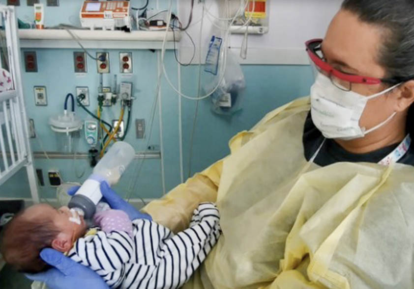 Healthcare worker in PPE bottle-feeding a baby in a hospital room with medical equipment