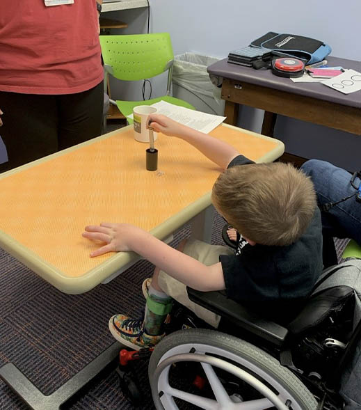Child in a wheelchair performing a fine motor task with a tool and object on a table. An adult is nearby
