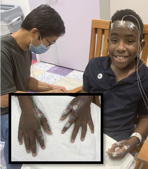 Smiling boy with electrodes on head and hands during testing. A masked healthcare worker assists. Inset: wired hands