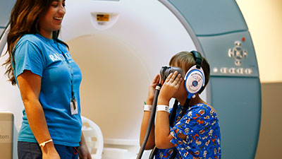 Child with headphones looks into a viewer near an MRI, supervised by smiling hospital staff