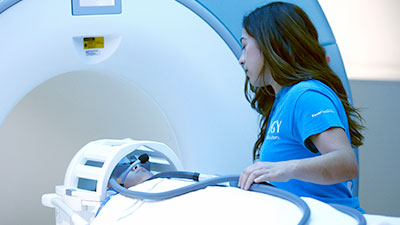 Patient in MRI machine, assisted by medical staff wearing blue 'OLOGY' shirt