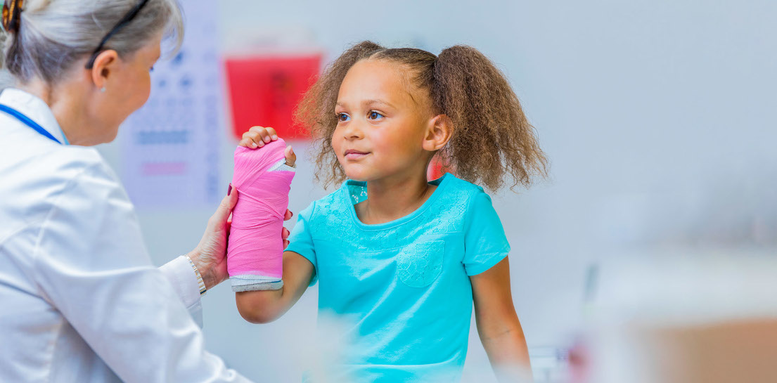 Pediatric orthopedic doctor examines an elementary age mixed race girl's broken arm. The girl has a pink cast on her arm.